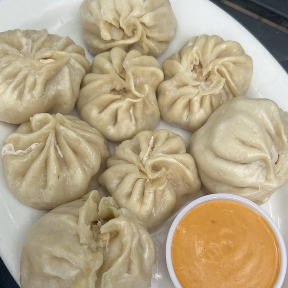 A plate of eight steamed Tibetan momos, with a small cup of orange dipping sauce.
