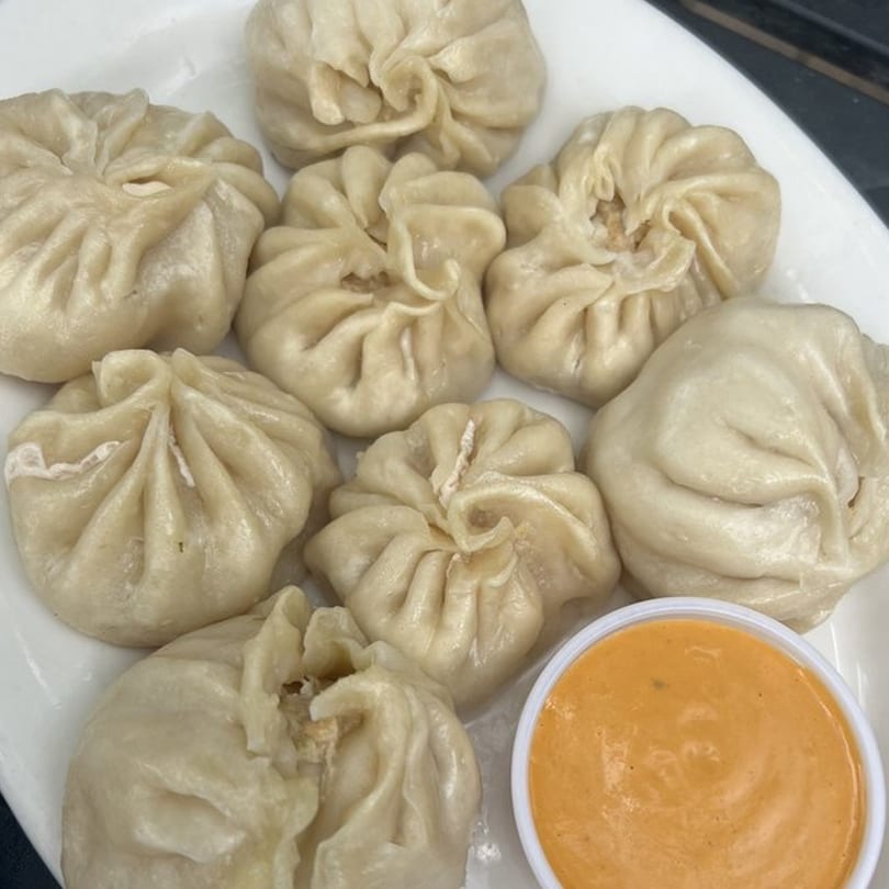 A plate of eight steamed Tibetan momos, with a small cup of orange dipping sauce.